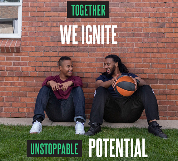 A male big and a male little sitting up against a brick wall smiling at each other while resting during their basketball game