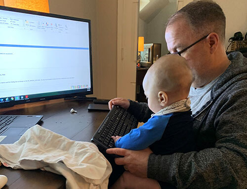 Man with glasses at a desk with a baby on his lap looking like the baby is learning how to use the computer