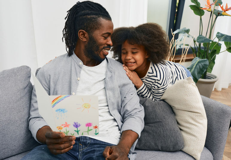 Father and daughter on a couch smiling at each other looking at a picture the daughter drew for the father