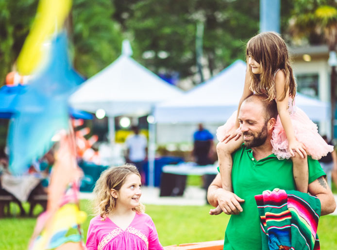 Father with two daughters one smiling to the right of him and the other on his shoulders at a festival