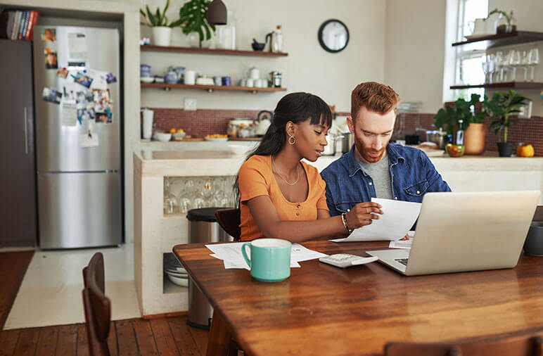 A young couple at their kitchen table looking at a papers in front of a laptop