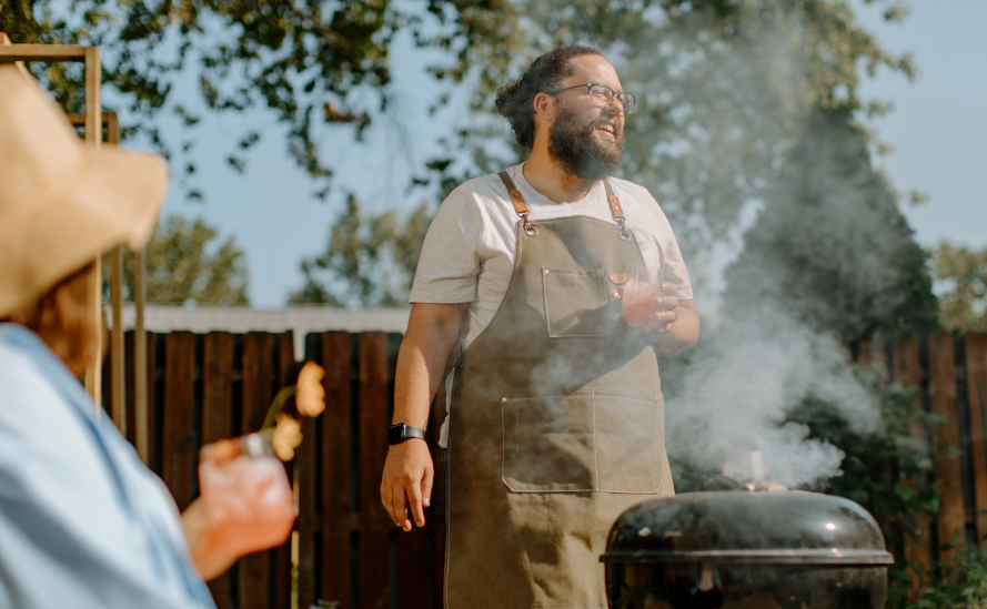 Bearded man enjoying an outdoor barbecue with a drink in hand, standing next to a grill on a sunny day, creating a relaxed backyard gathering vibe