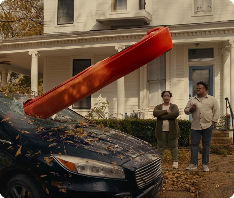 Damaged car with a large red object smashed through the windshield, parked in front of a house, with two people observing the scene